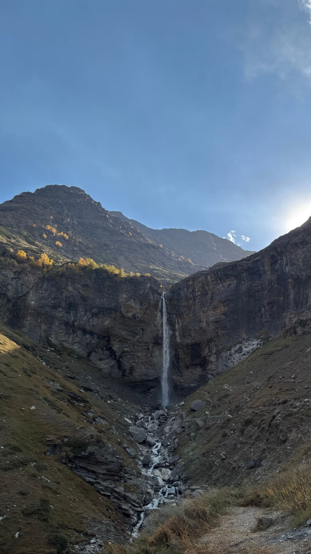 A thin waterfall dropping from a high cliff into a boulder-strewn ravine.