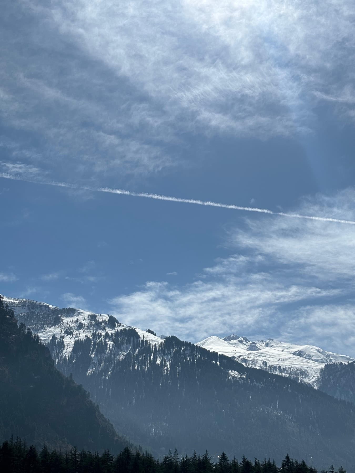 A contrail crossing the sky above a snow-dusted, forested ridgeline.