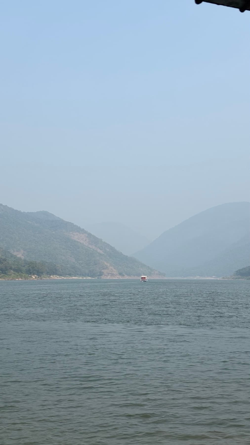 A lone boat on a wide river running between hazy forested hills.
