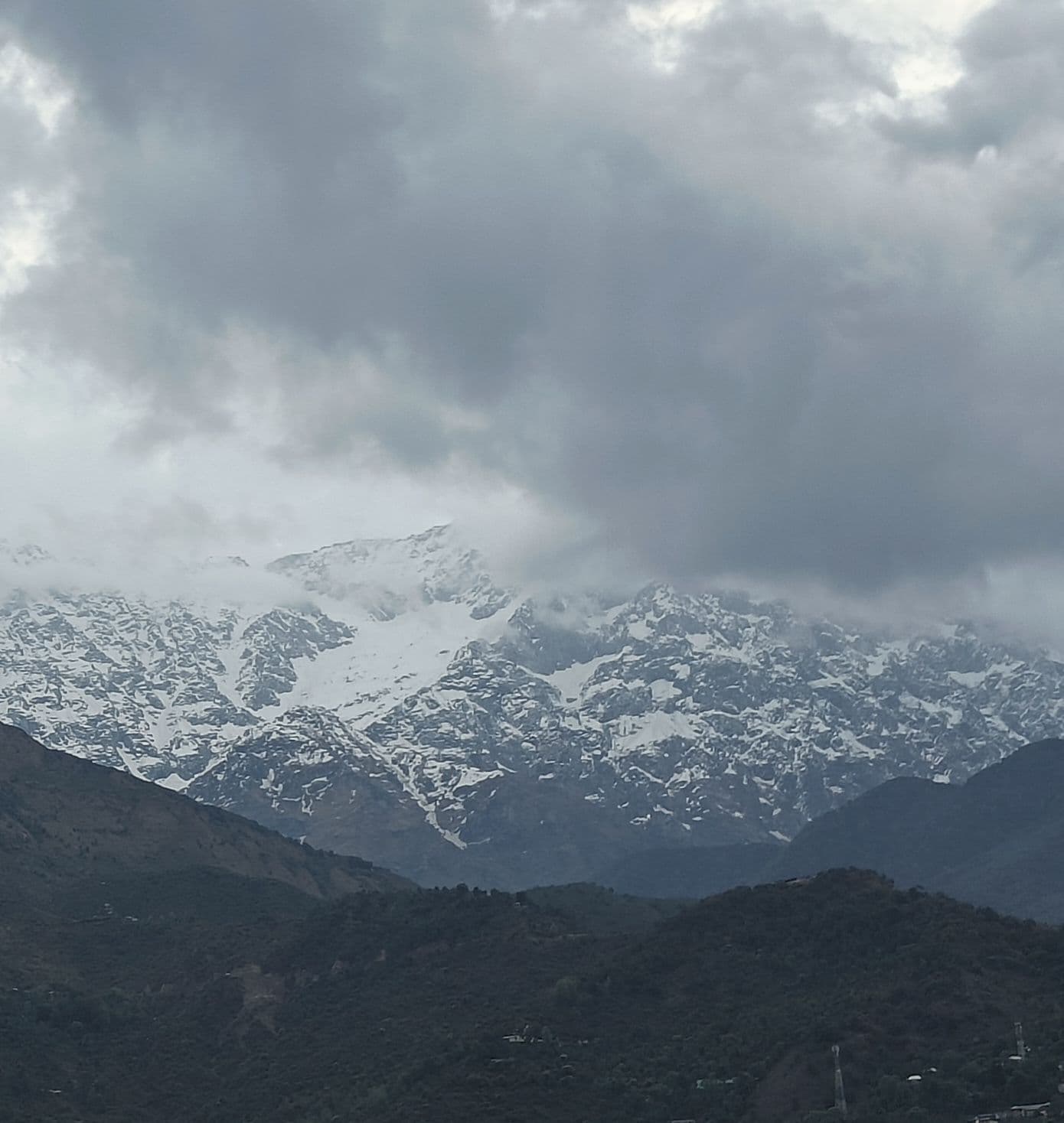 A snow-covered mountain range under heavy cloud, with a dark forested ridge in the foreground.