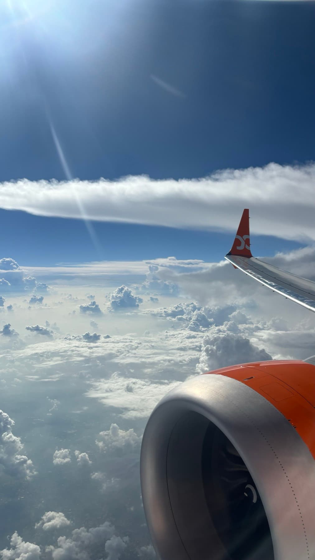 An aircraft wing and engine over a field of storm clouds, the sun flaring above.